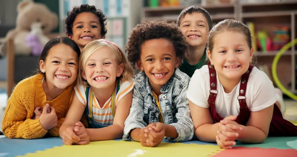 A group of six smiling children lie on colorful mats, facing the camera. They are in a playful and cheerful setting, with blurred toys and shelves in the background. The children show a mix of diverse appearances and joyful expressions.