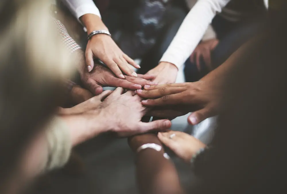 A group of diverse hands coming together in a unified stack, symbolizing teamwork and collaboration. The hands belong to individuals of different ages and ethnic backgrounds, highlighting inclusivity and solidarity.